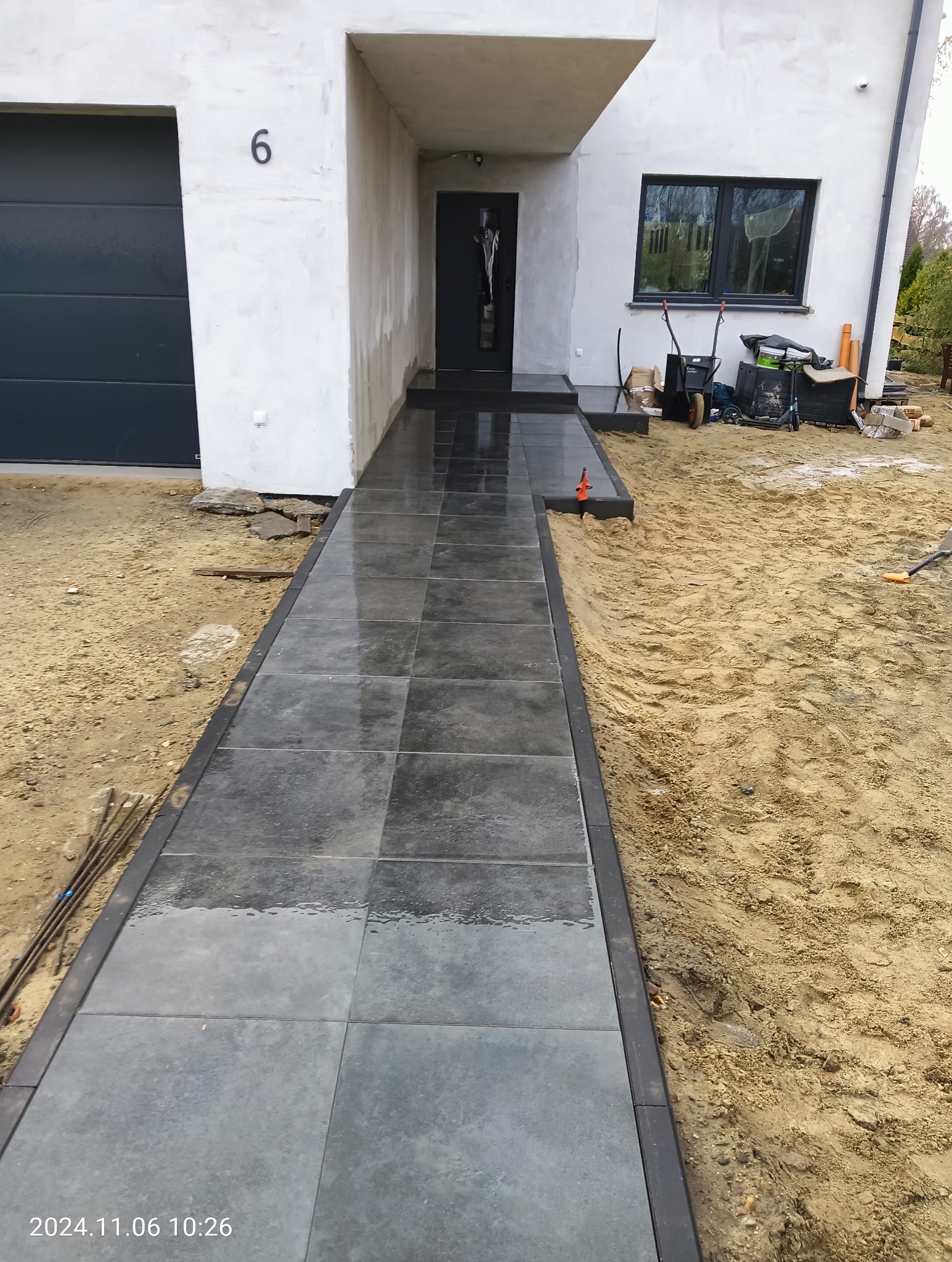 Świeżo ułożona, ciemnoszara ścieżka z kostki brukowej prowadząca do domu w Gdańsku, otoczona piaskiem i widocznymi narzędziami brukarskimi w tle.