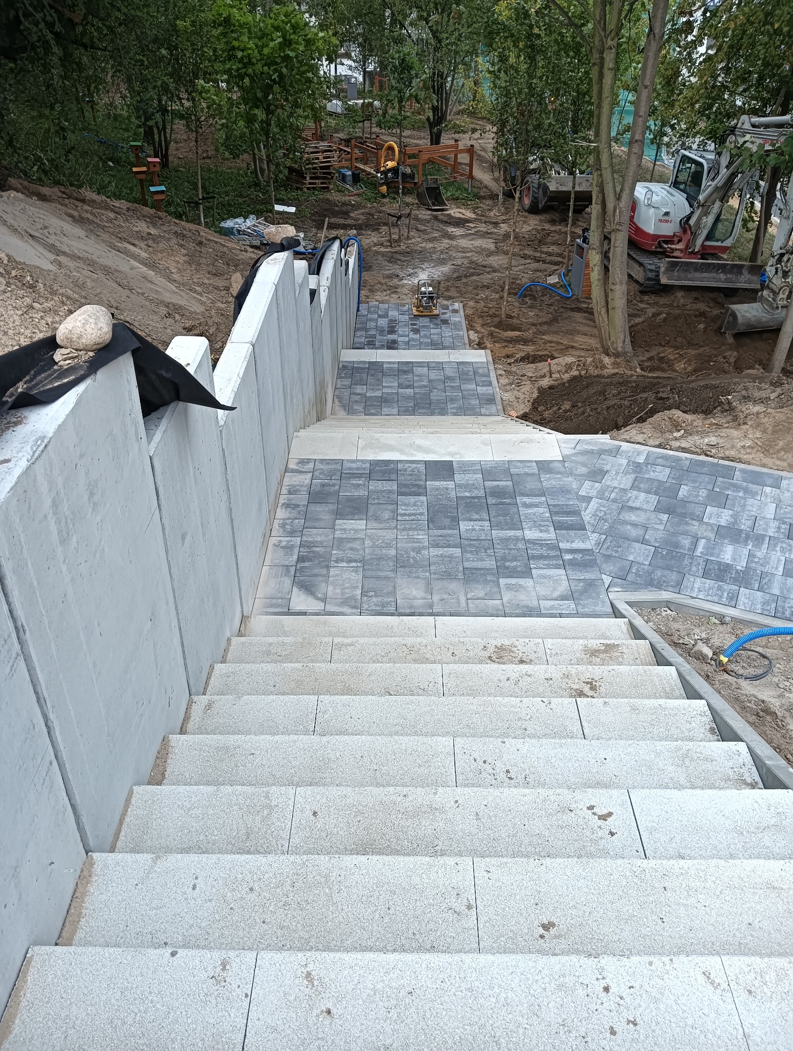 Schody z jasnych płyt prowadzą w dół, obok ściany oporowej i fragmentu ścieżki z szarej kostki; w tle widoczny teren budowy z mini koparką i ubijarką wibracyjną.