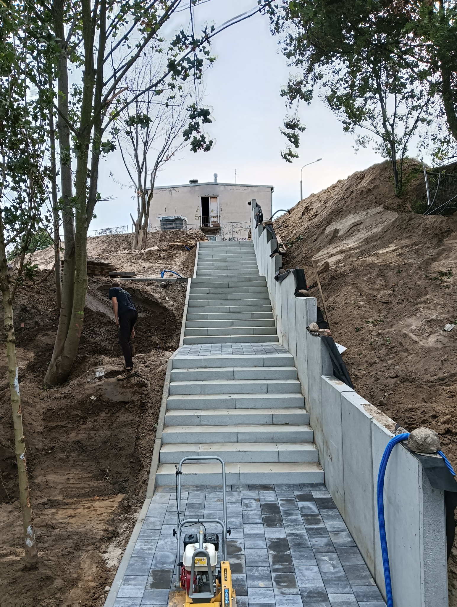 Schody z kostki brukowej prowadzące na wzgórze, z widoczną pracą ziemną po bokach i zagęszczarką w dolnej części kadru.