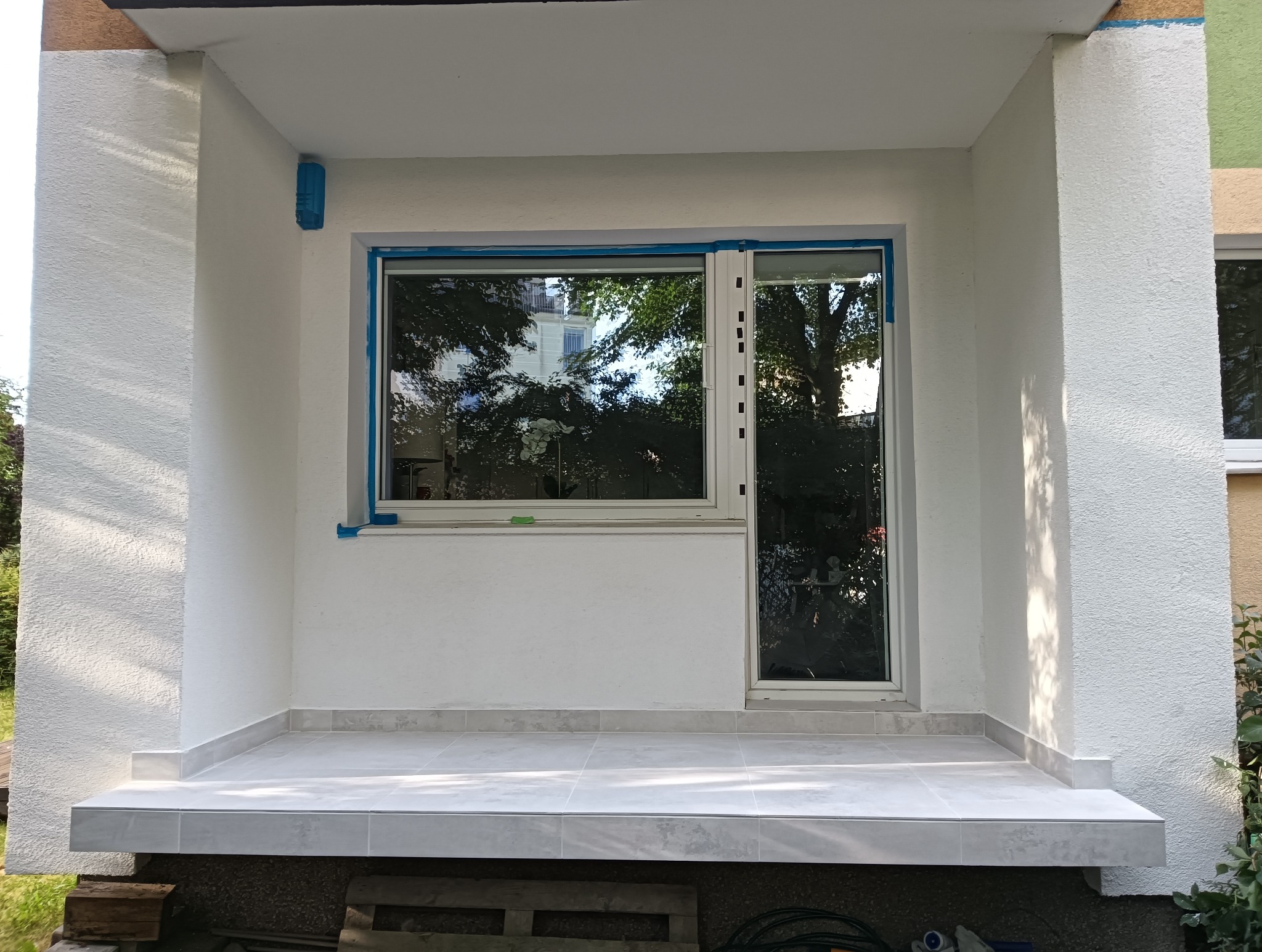 Wykończony balkon z białymi ścianami o chropowatej strukturze, szarymi płytkami na podłodze i nowymi oknami zabezpieczonymi taśmą malarską.