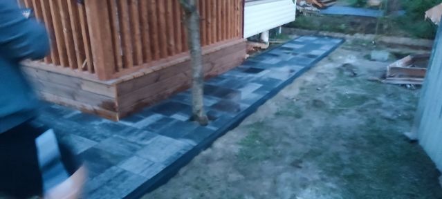 Ścieżka z szarej kostki brukowej prowadząca od drewnianego tarasu wzdłuż ściany budynku na zewnątrz, obok rosnącego drzewa.