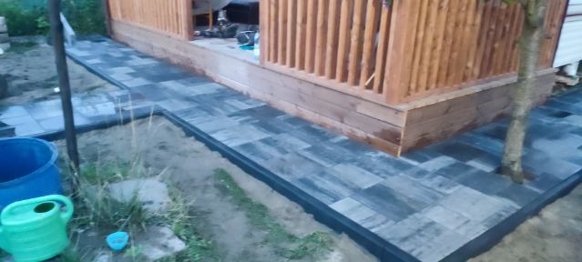 Ułożona kostka brukowa w kolorze szaro-czarnym, tworząca ścieżkę wokół drewnianej altany, z widocznym niebieskim wiadrem i zieloną konewką w tle.