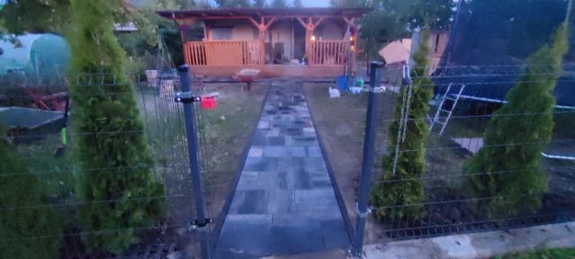 Nowo ułożona ścieżka z szarej kostki brukowej prowadząca do drewnianej altany, widziana przez otwartą, metalową furtkę ogrodzenia obsadzoną tujami.