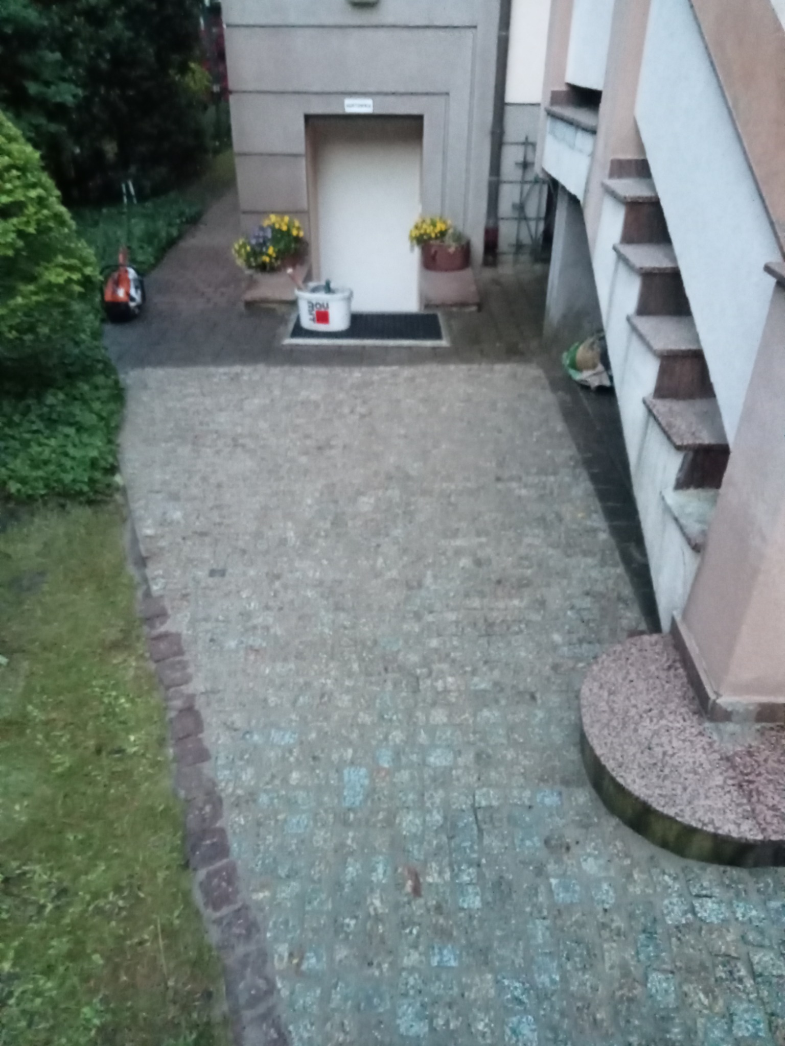 Ułożona kostka brukowa na podjeździe przed budynkiem, widoczny trawnik, schody i pojemnik po farbie.