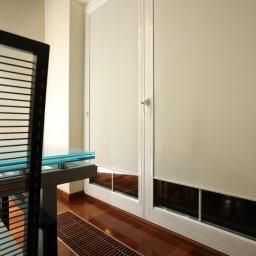 Sun & Glass Technik Sp. z o.o. - Dwa okna z białymi ramami i opuszczonymi, kremowymi roletami wewnętrznymi, widoczne kratki wentylacyjne w podłodze i fragment nowoczesnego krzesła z przezroczystym blatem stołu.
