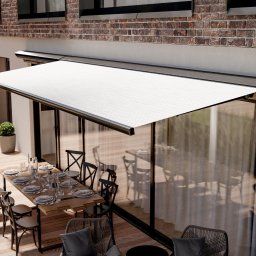 Sun & Glass Technik Sp. z o.o. - Elegancka, rozłożona markiza w kolorze białym nad tarasem z dużym oknem, na którym widać stół z zastawą dla wielu osób, ceglana ściana budynku w tle.