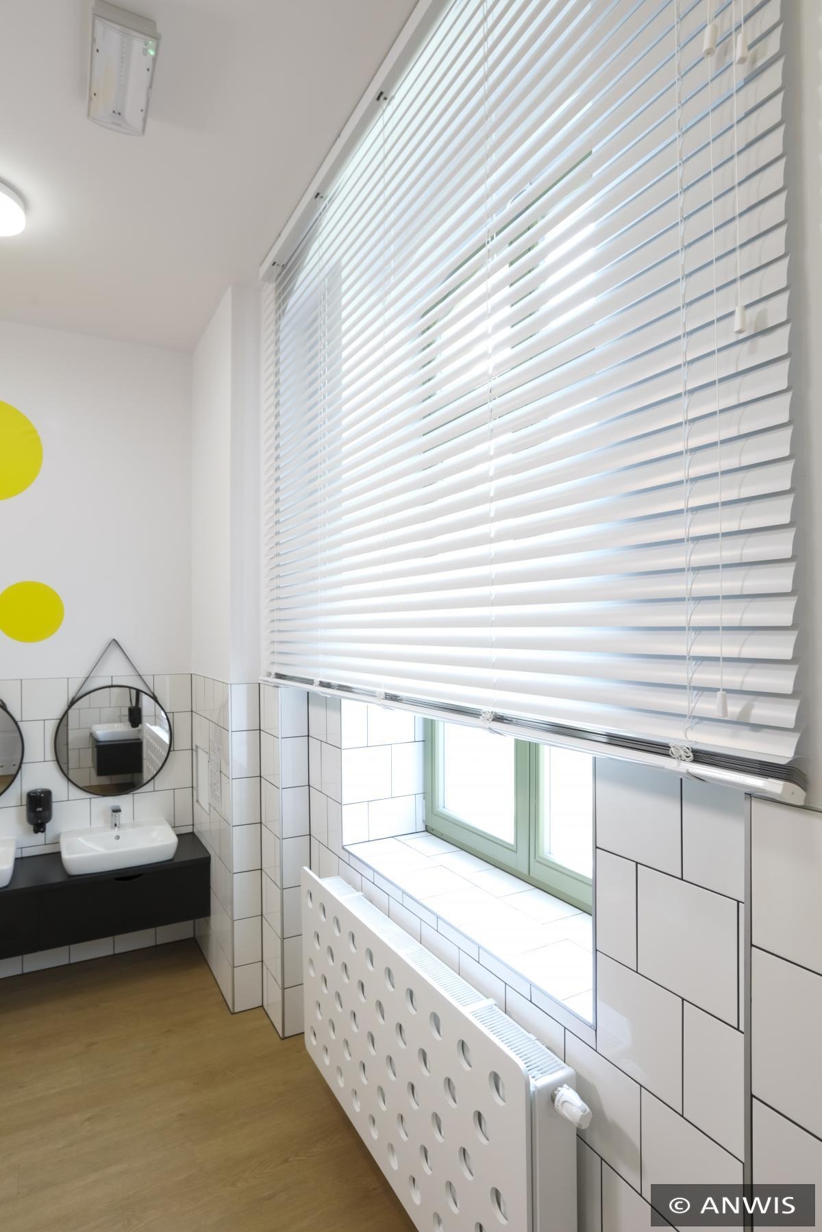 Białe żaluzje aluminiowe zamontowane w oknie łazienki z białymi płytkami i grzejnikiem z perforacją, widok z wnętrza.