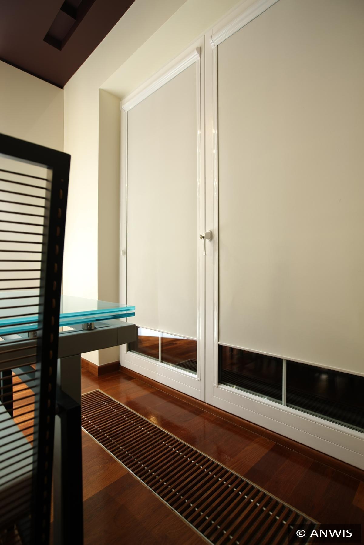 Dwa okna z białymi ramami i opuszczonymi, kremowymi roletami wewnętrznymi, widoczne kratki wentylacyjne w podłodze i fragment nowoczesnego krzesła z przezroczystym blatem stołu.