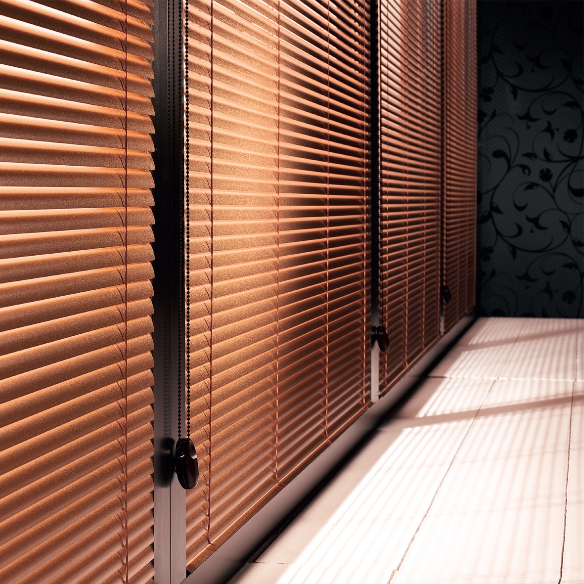 Pomarańczowe żaluzje z łańcuszkowym mechanizmem regulacji, zamontowane w pomieszczeniu z ciemną tapetą w kwiatowe wzory i jasną podłogą.