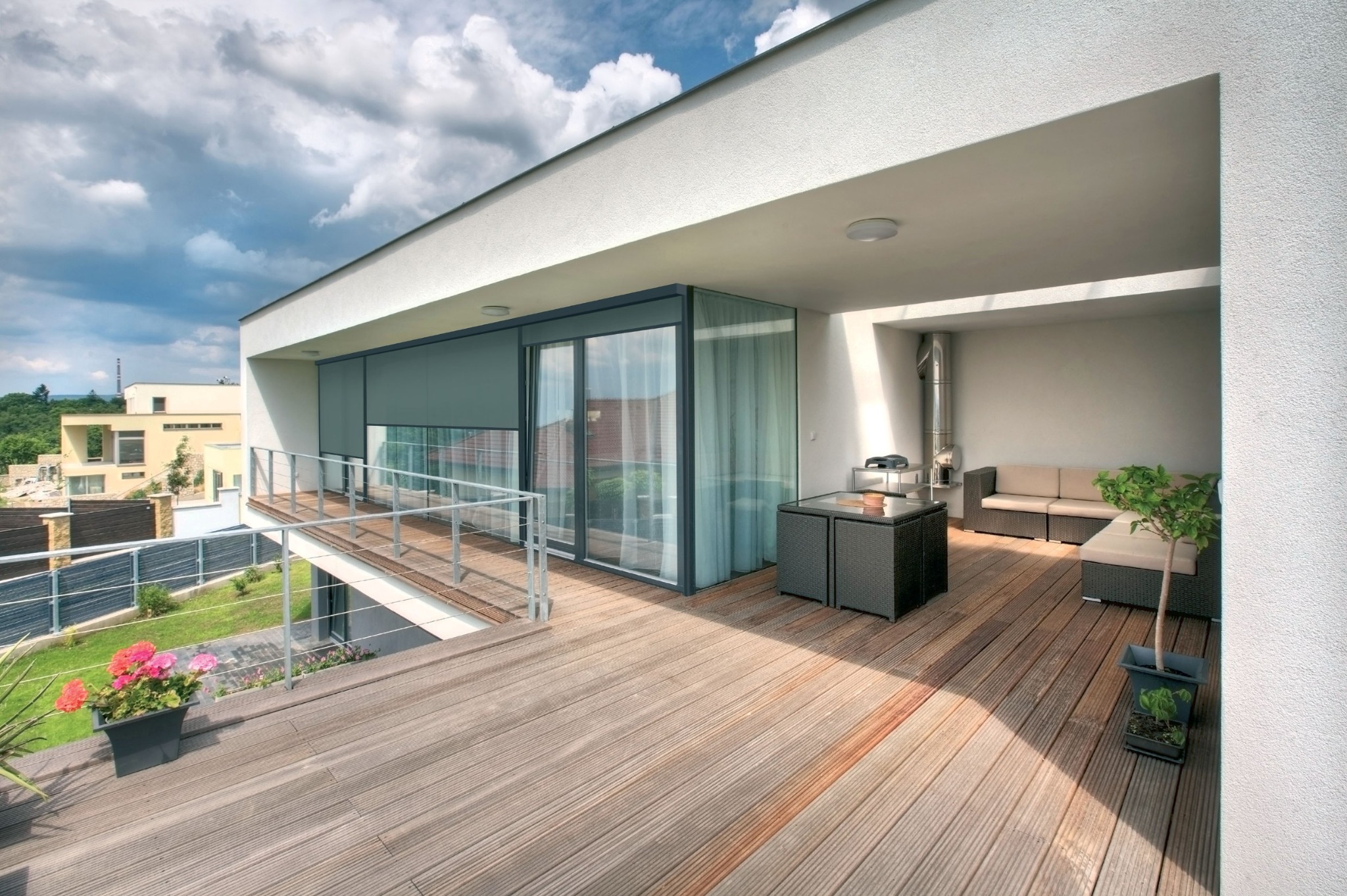 Nowoczesny taras z drewnianą podłogą, osłonięty szarą tkaniną typu screen, chroniącą przed słońcem, z widokiem na ogród i sąsiednie budynki.