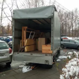 Otwarta paka samochodu dostawczego wypełniona meblami i przedmiotami podczas zimowej przeprowadzki, widoczne zaparkowane samochody i śnieg na chodniku.