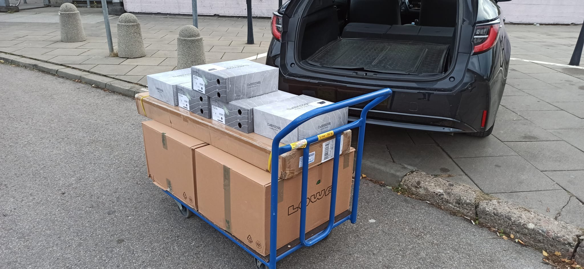Niebieski wózek transportowy załadowany kartonami i pudełkami z butami marki LOWA, stojący przed otwartym bagażnikiem czarnego samochodu osobowego.
