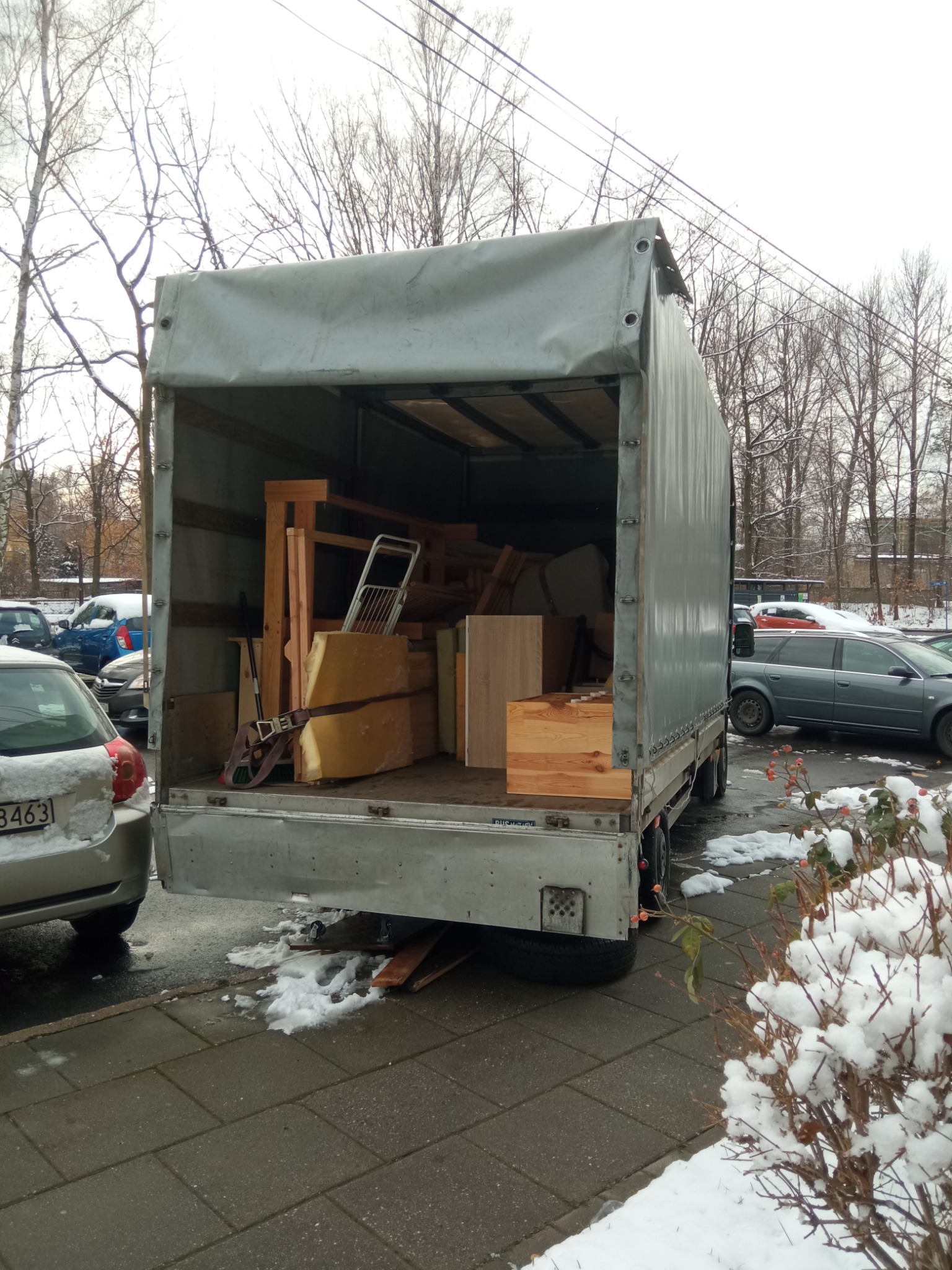 Otwarta paka samochodu dostawczego wypełniona meblami i przedmiotami podczas zimowej przeprowadzki, widoczne zaparkowane samochody i śnieg na chodniku.
