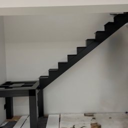 Dominik Gawarecki - Czarne schody metalowe bez poręczy, z widoczną konstrukcją podestu, w surowym wnętrzu. Na podłodze płyty i narzędzia, co sugeruje etap montażu.