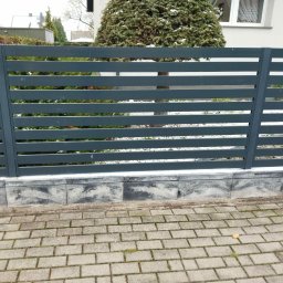 Dominik Gawarecki - Nowoczesne, poziome ogrodzenie panelowe w kolorze grafitowym, osadzone na betonowej podmurówce. W tle widoczne krzewy i fragment budynku.