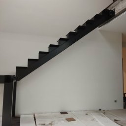 Dominik Gawarecki - Nowoczesne, minimalistyczne schody metalowe w kolorze czarnym, bez poręczy, zamontowane przy białej ścianie. Widoczne zabezpieczenia podłogi na czas montażu.