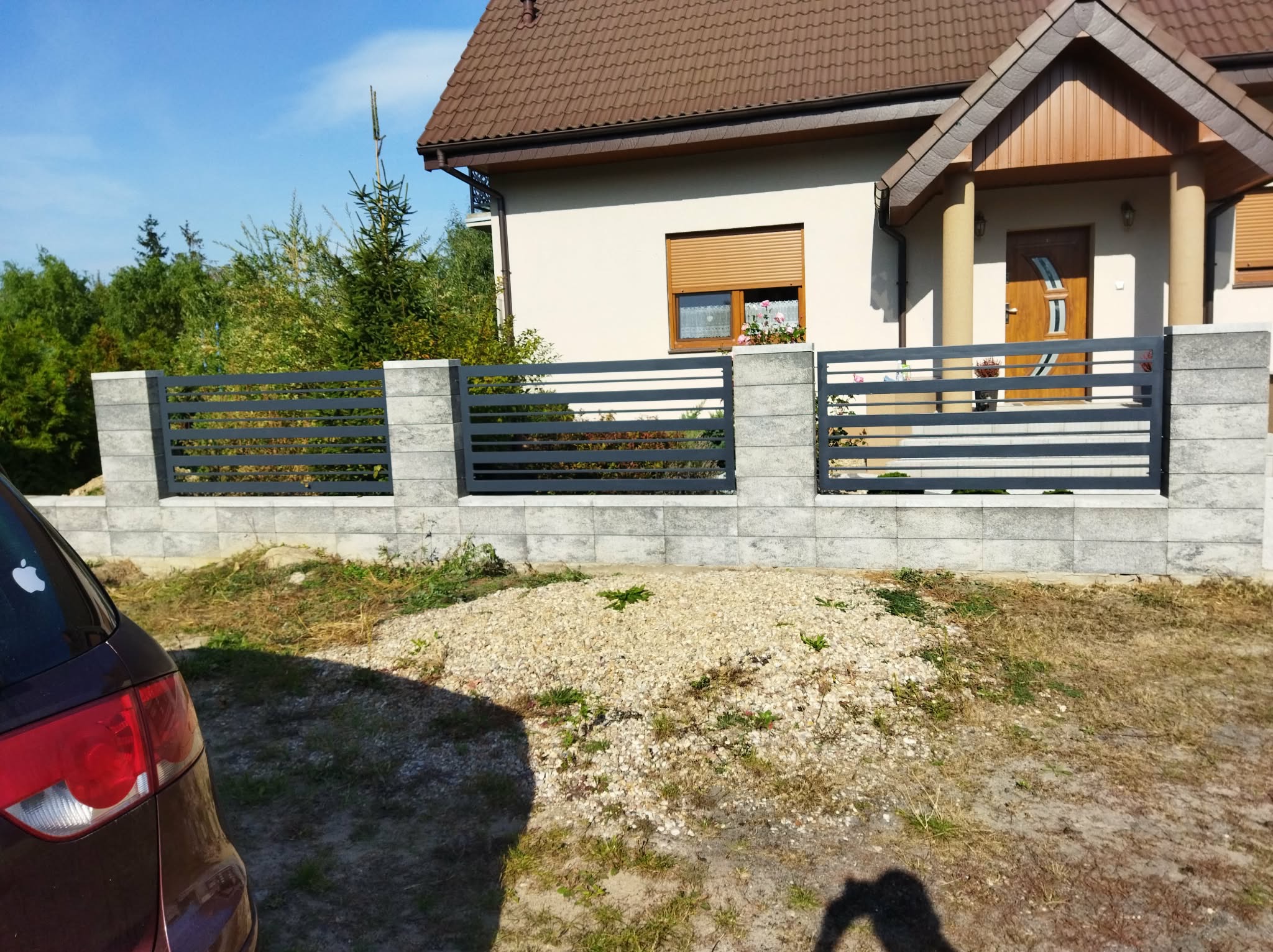 Nowoczesne ogrodzenie panelowe z poziomymi przęsłami w kolorze grafitowym, osadzone na betonowych słupkach i podmurówce, na tle domu jednorodzinnego w słoneczny dzień.