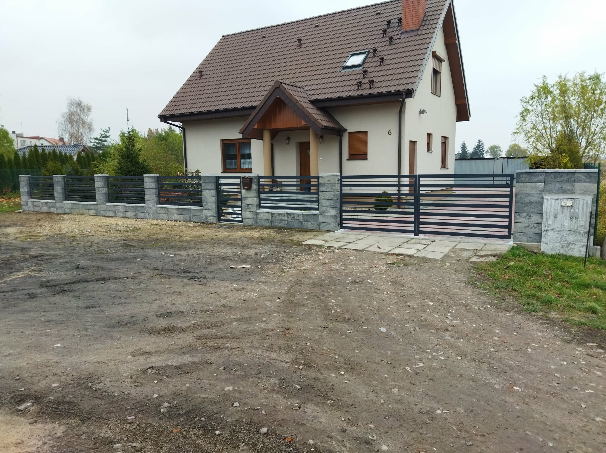 Nowoczesne ogrodzenie posesji z przesuwną bramą i segmentami z poziomych, metalowych profili, osadzone na słupkach z szarej kostki. Dom jednorodzinny w tle.
