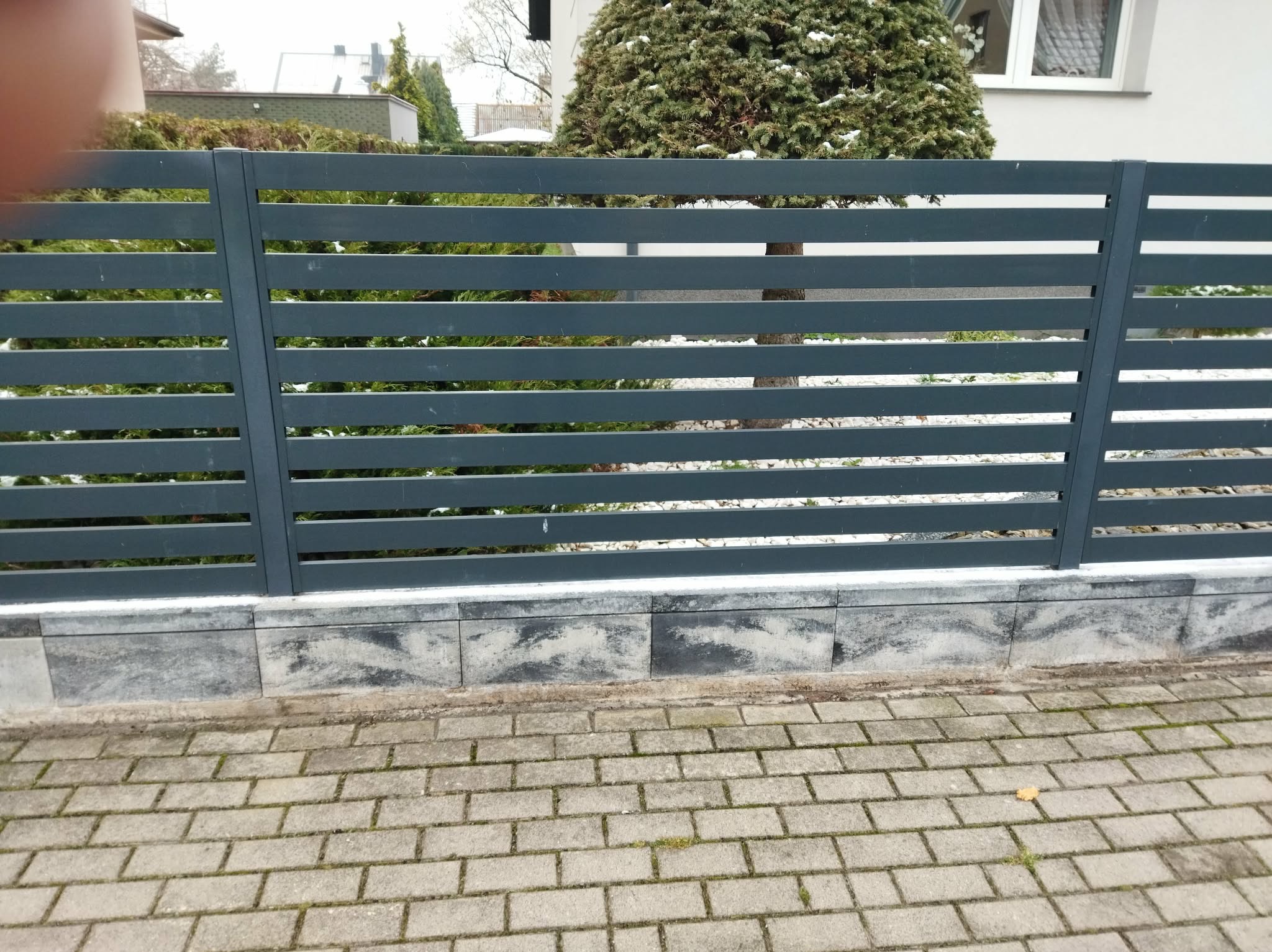 Nowoczesne, poziome ogrodzenie z metalowych profili w kolorze grafitowym, osadzone na betonowej podmurówce z kostki. W tle zieleń ogrodu i fragment budynku.