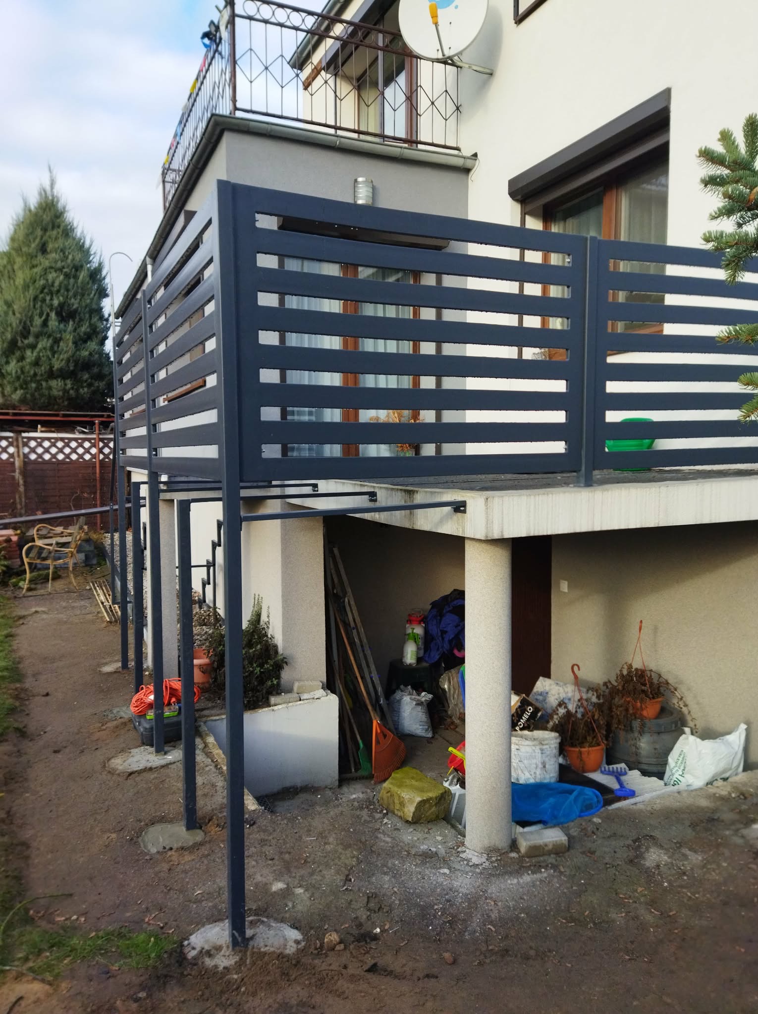 Nowoczesna, pozioma balustrada balkonowa z metalu w kolorze grafitowym, zamontowana na betonowym balkonie. Widok z zewnątrz, z widocznymi podporami i otoczeniem.