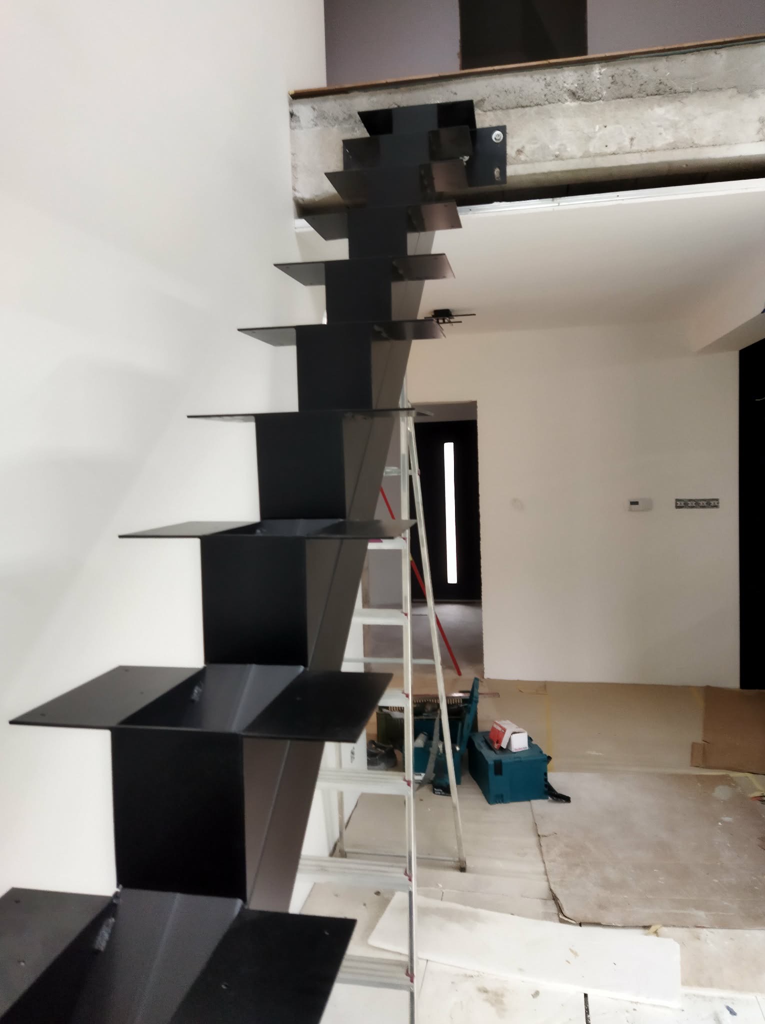 Nowoczesne, czarne schody metalowe wspornikowe w trakcie montażu. Widoczna drabina i narzędzia. Minimalistyczny design, surowe wnętrze w trakcie wykończenia.