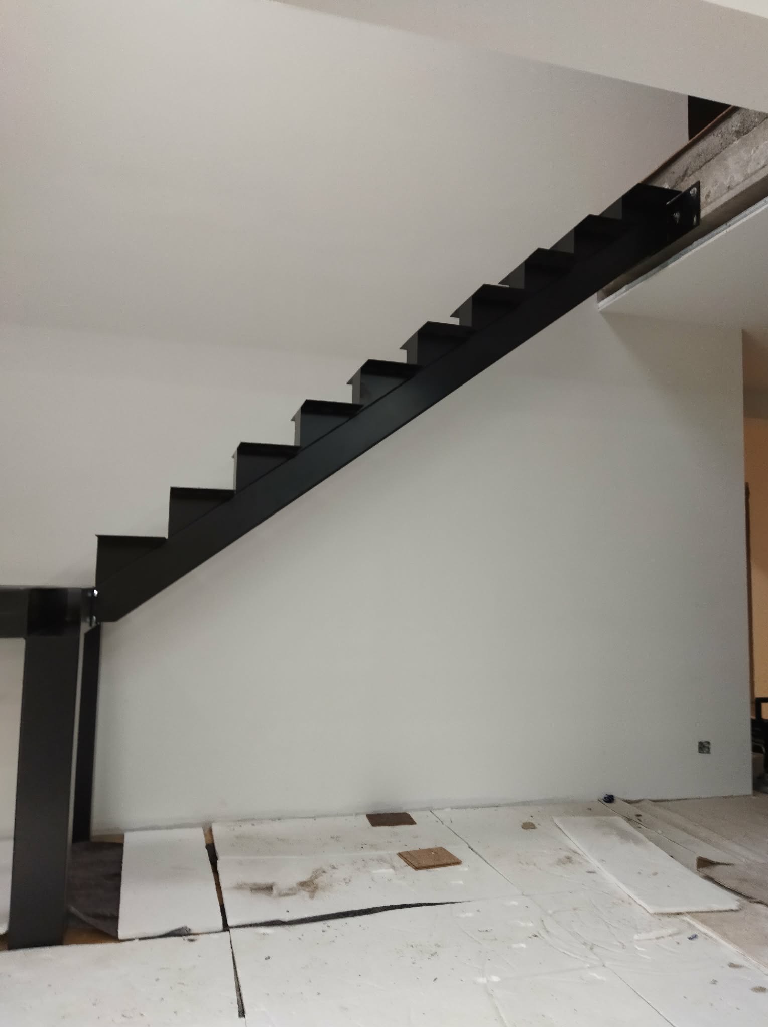 Nowoczesne, minimalistyczne schody metalowe w kolorze czarnym, bez poręczy, zamontowane przy białej ścianie. Widoczne zabezpieczenia podłogi na czas montażu.