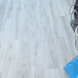 Świeżo ułożona, jasnoszara podłoga z paneli winylowych imitujących drewno, widoczny odkurzacz przemysłowy i niebieski worek na śmieci w rogu pomieszczenia.