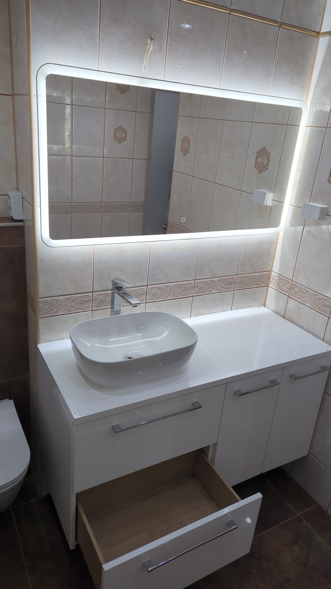 Biała umywalka nablatowa z chromowaną baterią na szafce z szufladami w łazience z beżowymi płytkami, podświetlane lustro LED, otwarta szuflada.