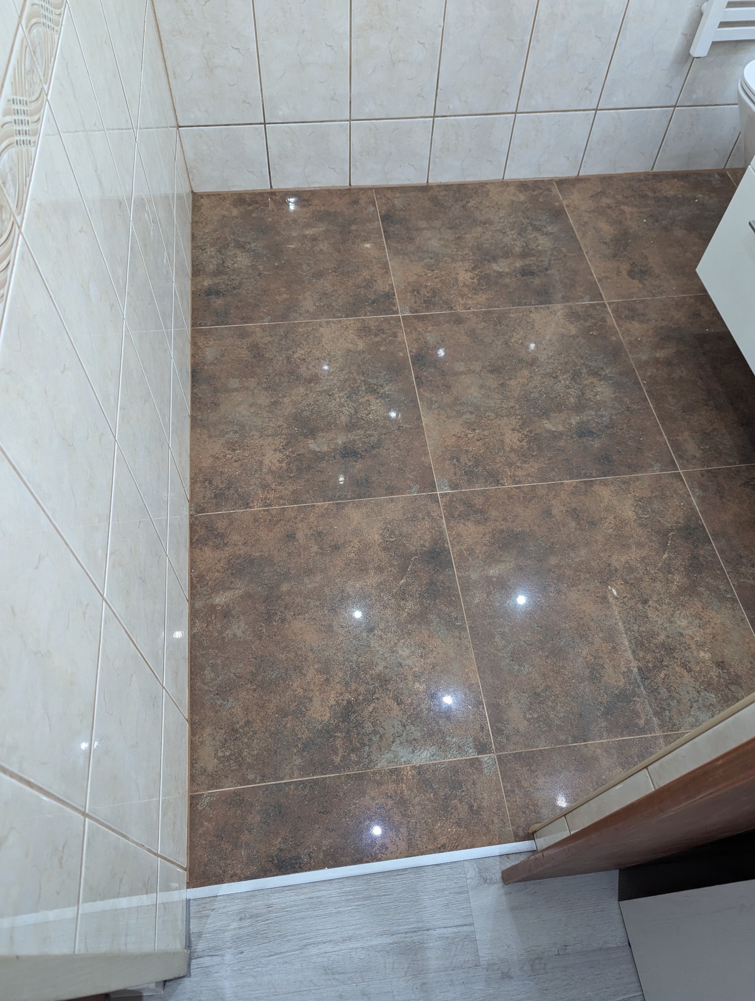 Brązowe płytki podłogowe w łazience, kremowe płytki ścienne, widoczna listwa przypodłogowa i fragment białej armatury łazienkowej.