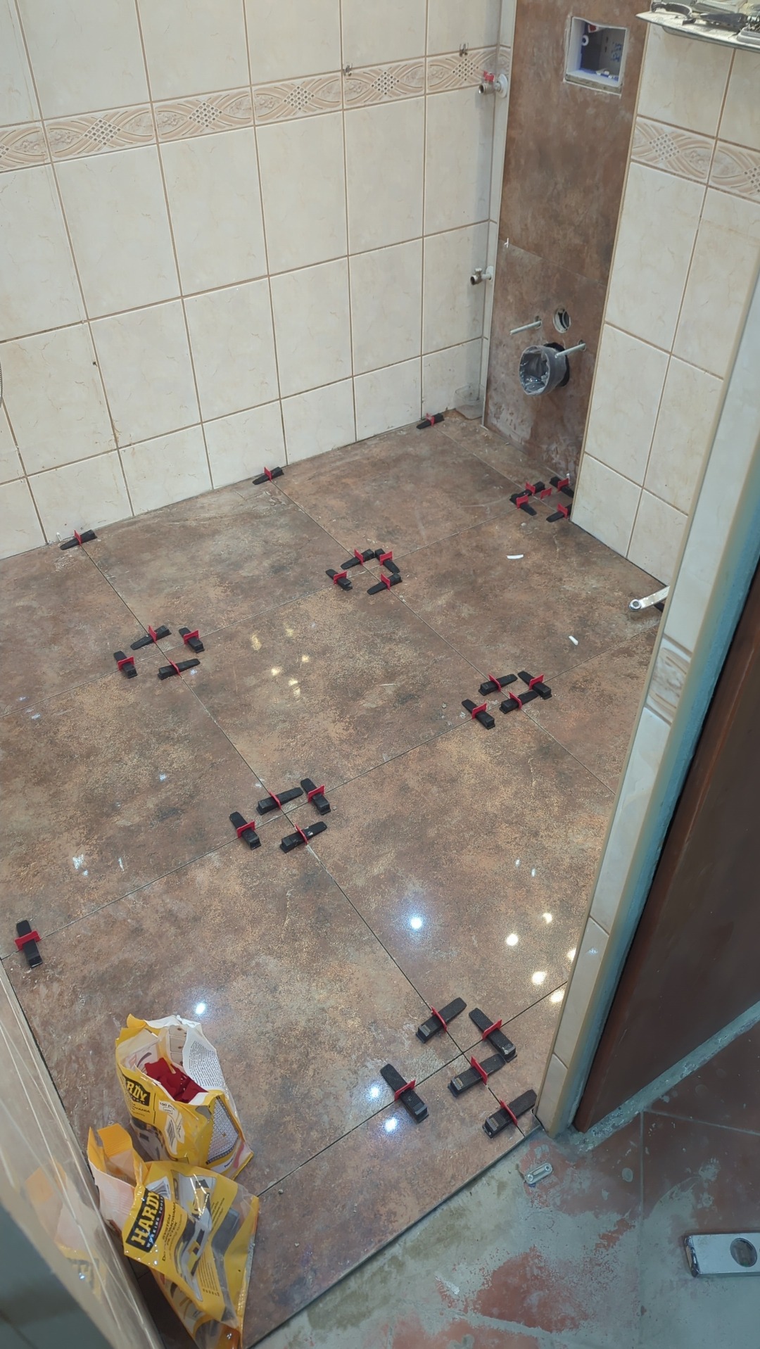 Brązowe płytki podłogowe w trakcie układania w łazience, widoczne plastikowe krzyżyki dystansowe i opakowania po kleju.