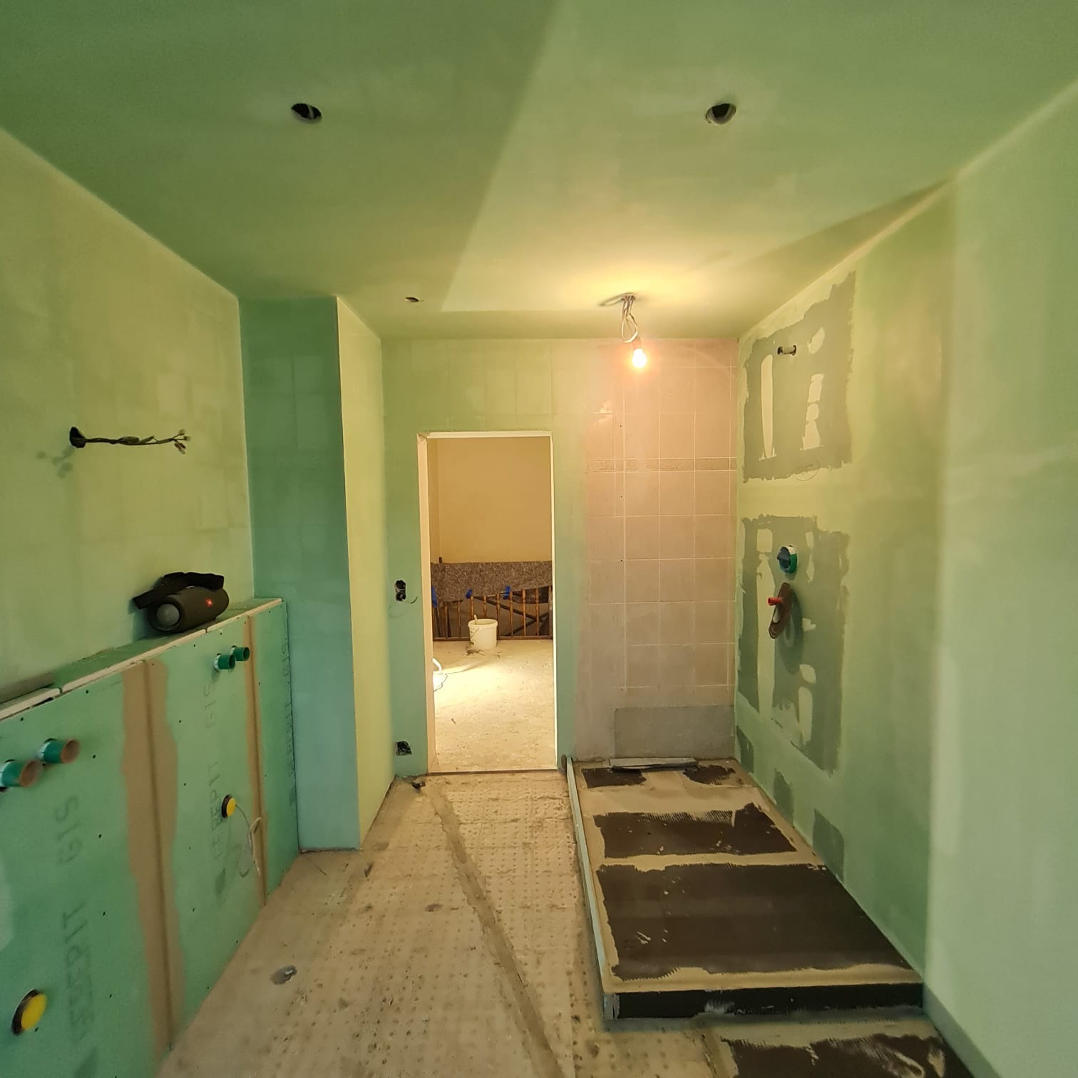 Wnętrze łazienki w trakcie prac wykończeniowych: zielone płyty gipsowo-kartonowe na ścianach, częściowo położone płytki, przygotowany brodzik, widoczne instalacje, surowe ściany z widocznymi...