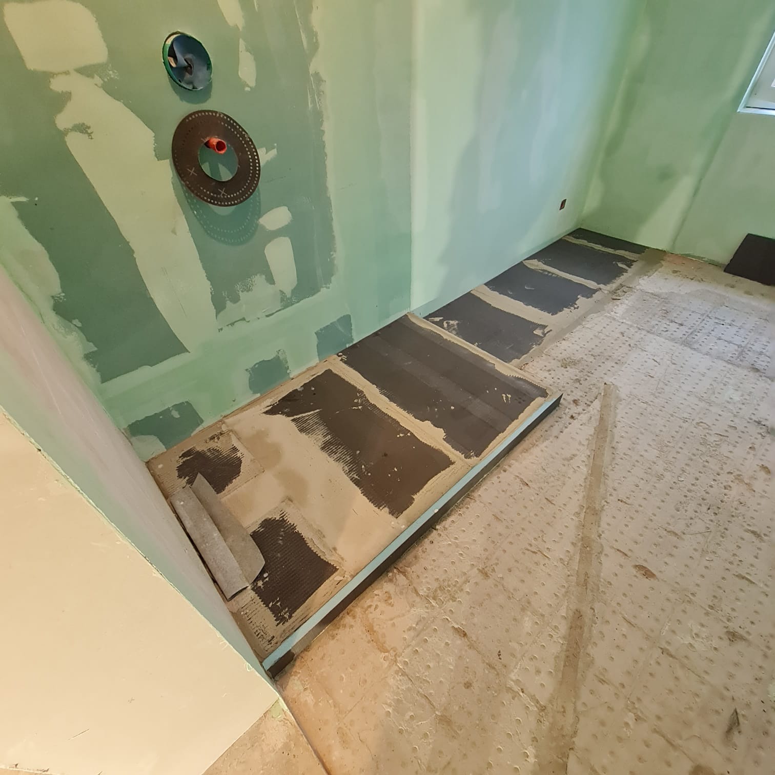 Wnętrze łazienki w trakcie prac wykończeniowych; ściany pokryte zielonymi płytami gipsowo-kartonowymi z widocznymi śladami szpachlowania, podłoga częściowo przygotowana pod płytki, widoczne...