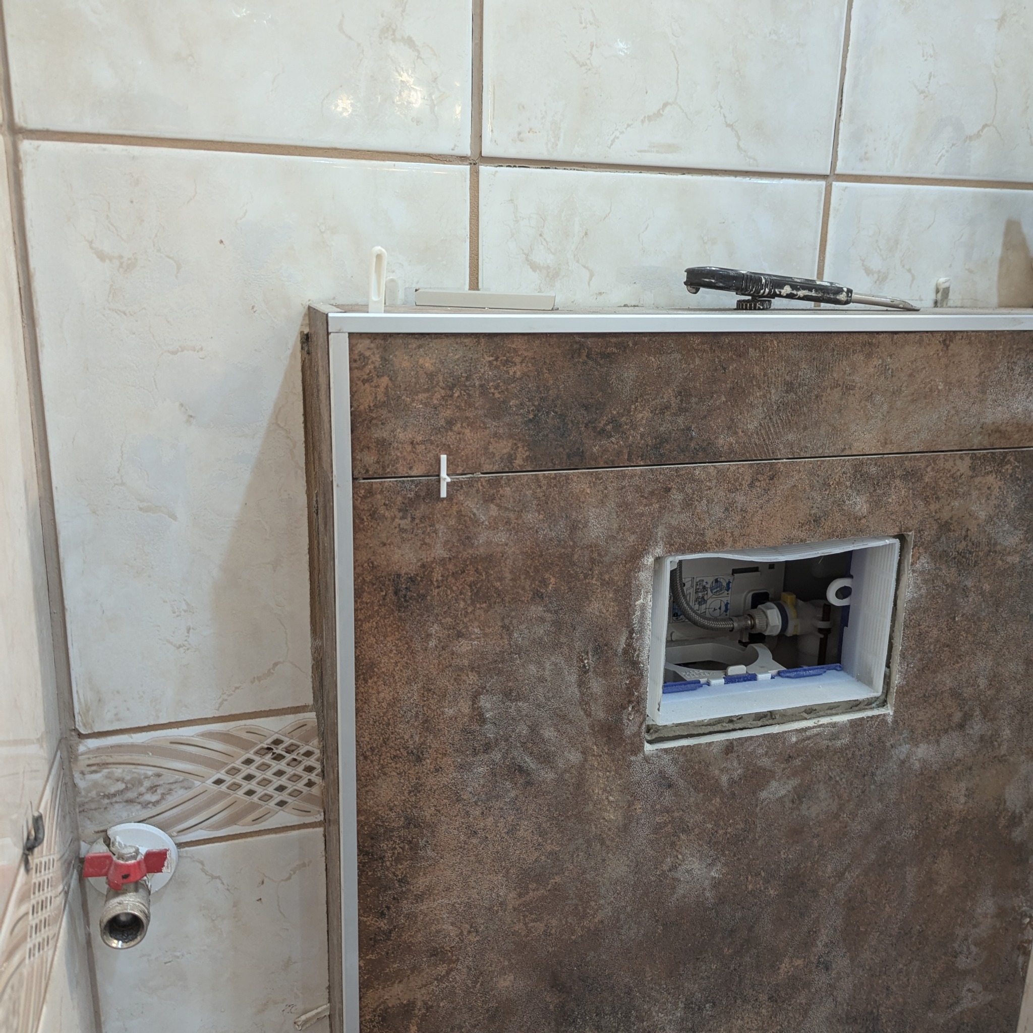 Podtynkowa instalacja WC w trakcie montażu, widoczne płytki imitujące kamień, zawór odcinający wodę i narzędzia na obudowie.