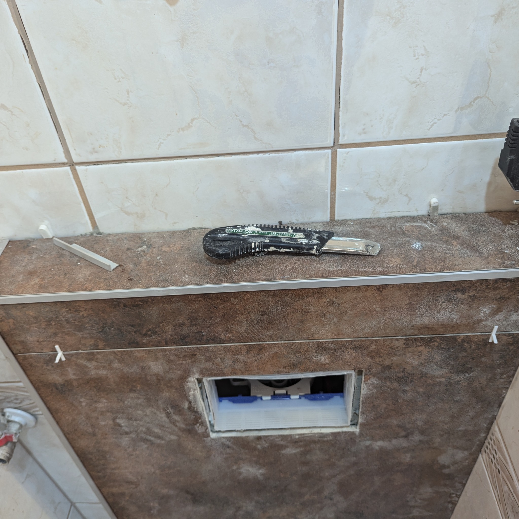 Montaż brązowych płytek w łazience, widoczny otwór na stelaż podtynkowy, listwa wykończeniowa, krzyżyki dystansowe i zabrudzony nożyk do cięcia płytek marki Stalco.