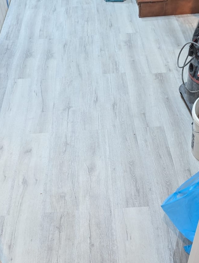 Świeżo ułożona, jasnoszara podłoga z paneli winylowych imitujących drewno, widoczny odkurzacz przemysłowy i niebieski worek na śmieci w rogu pomieszczenia.