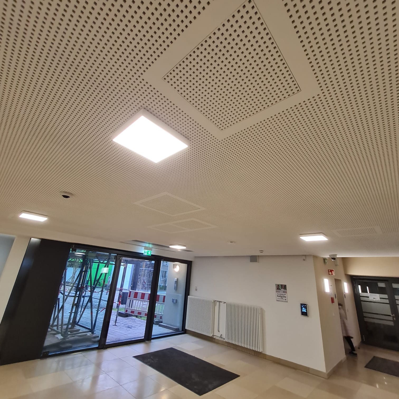 Sufit podwieszany z perforacją kwadratową i wbudowanym oświetleniem LED w holu budynku z widocznymi drzwiami wejściowymi i grzejnikami.