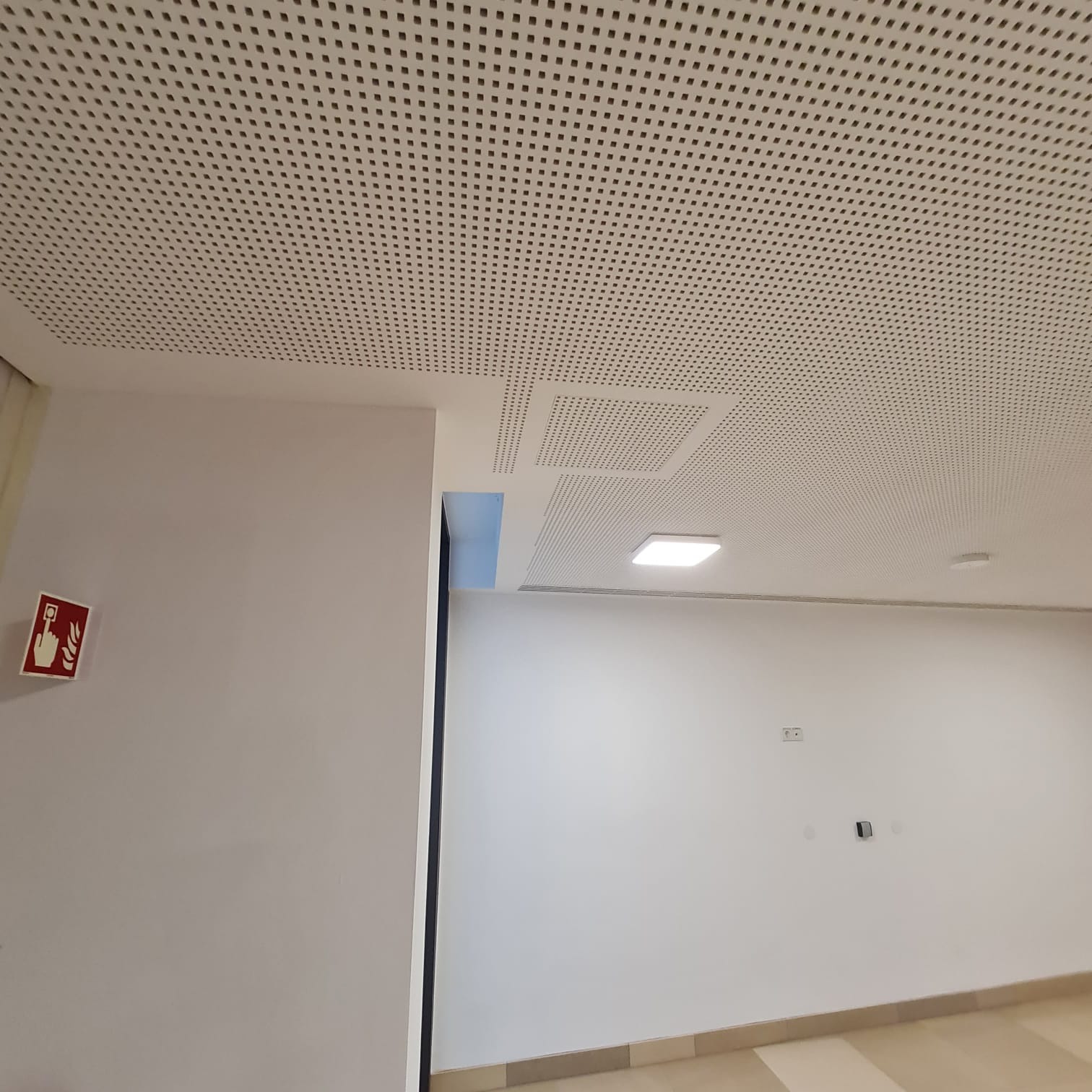 Perforowany sufit podwieszany z kwadratowymi otworami, widok z dołu, białe ściany i znak ewakuacyjny