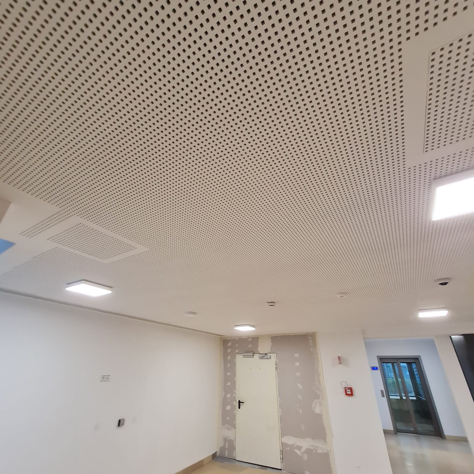 Sufit podwieszany z perforacją kwadratową, widoczne panele oświetleniowe LED i czujniki dymu, ściany w trakcie przygotowania do malowania, widoczny fragment windy.