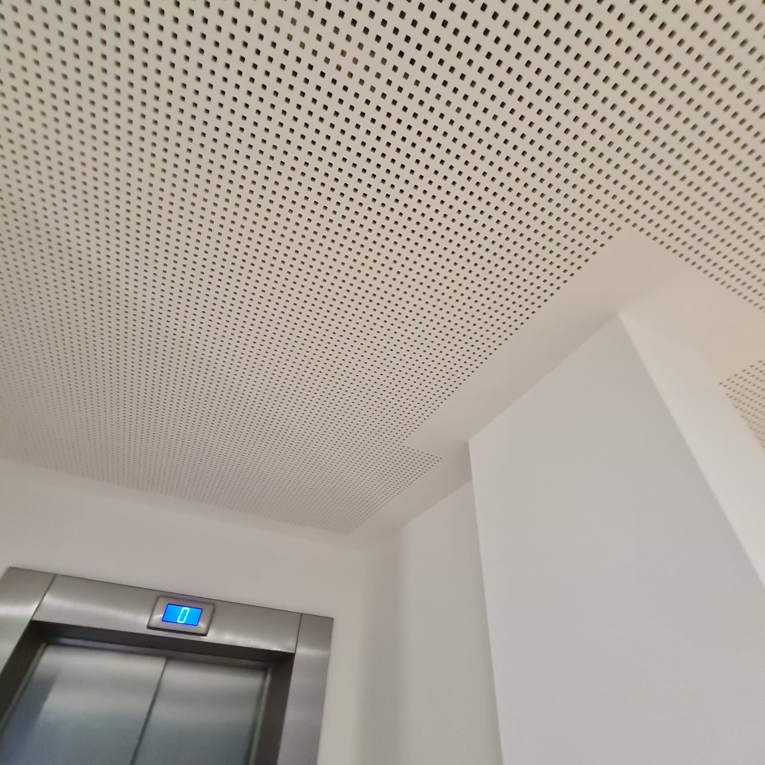 Perforowany sufit podwieszany z kwadratowymi otworami, widoczny fragment windy z wyświetlaczem cyfrowym wskazującym zero, białe ściany w tle.