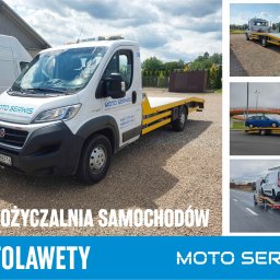 Moto Serwis - Biała laweta z żółtymi elementami zabezpieczającymi, zaparkowana na żwirowym podłożu, z logo 'Moto Serwis' i numerami kontaktowymi na kabinie, w tle inne pojazdy i zieleń.