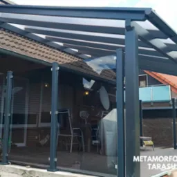 Zadaszony taras z czarną konstrukcją, szklanymi panelami i widocznym wyposażeniem: stół, krzesła i grill przykryty pokrowcem.