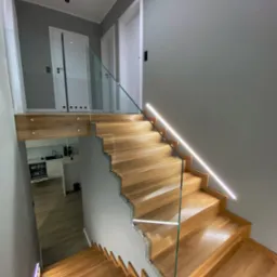 Nowoczesne schody z drewna z podświetleniem LED wzdłuż ściany i szklaną balustradą, widok z góry na dół, w tle widoczny fragment kuchni.