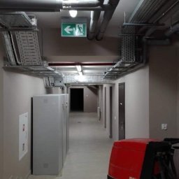 Star Gold - Długi korytarz biurowy z szarymi ścianami i podłogą z płytek, oświetlony jarzeniówkami, z widocznymi instalacjami pod sufitem i czerwoną maszyną do czyszczenia podłóg w prawym dolnym rogu.