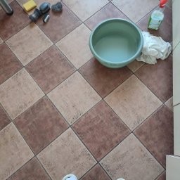 Star Gold - Ceramiczna podłoga w brązowo-beżową szachownicę z widocznymi śladami czyszczenia, miska z wodą, butelka ze spryskiwaczem, ściereczka, gąbka, szczotki i stopy osoby w białych klapkach.