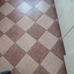 Star Gold - Brązowo-beżowa podłoga z płytek ułożonych w karo, widoczne białe klapki i fragment narzędzia do fugowania przy krawędzi, szafka kuchenna w tle.