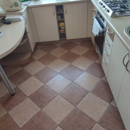 Star Gold - Kuchnia z brązowo-beżową podłogą w szachownicę, jasnymi szafkami i blatem, widok na okno z roślinami.