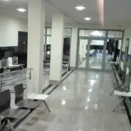 Wnętrze poczekalni z rzędem metalowych krzeseł, marmurową podłogą i białymi kolumnami, widok na szklane drzwi wejściowe.