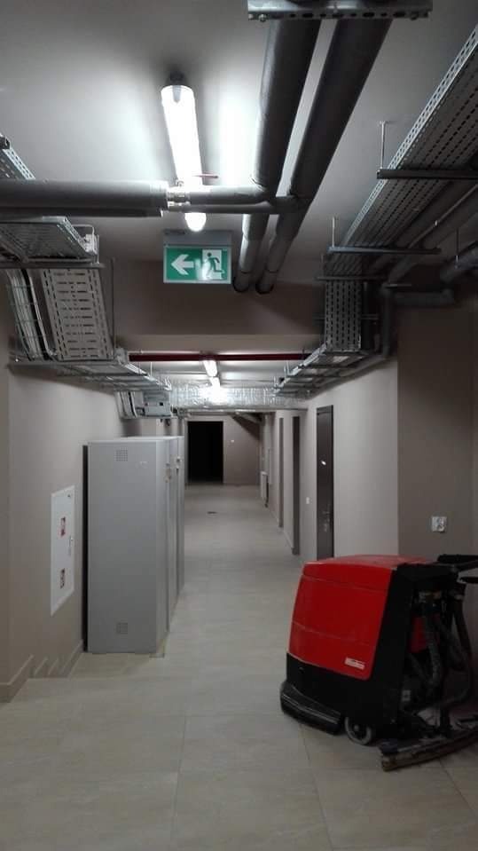 Długi korytarz biurowy z szarymi ścianami i podłogą z płytek, oświetlony jarzeniówkami, z widocznymi instalacjami pod sufitem i czerwoną maszyną do czyszczenia podłóg w prawym dolnym rogu.