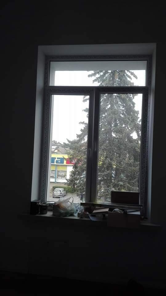 Białe, plastikowe okno z widokiem na ulicę, drzewo iglaste i budynek sklepowy; na parapecie widoczne ślady po remoncie, takie jak folie, puszki i kartony.