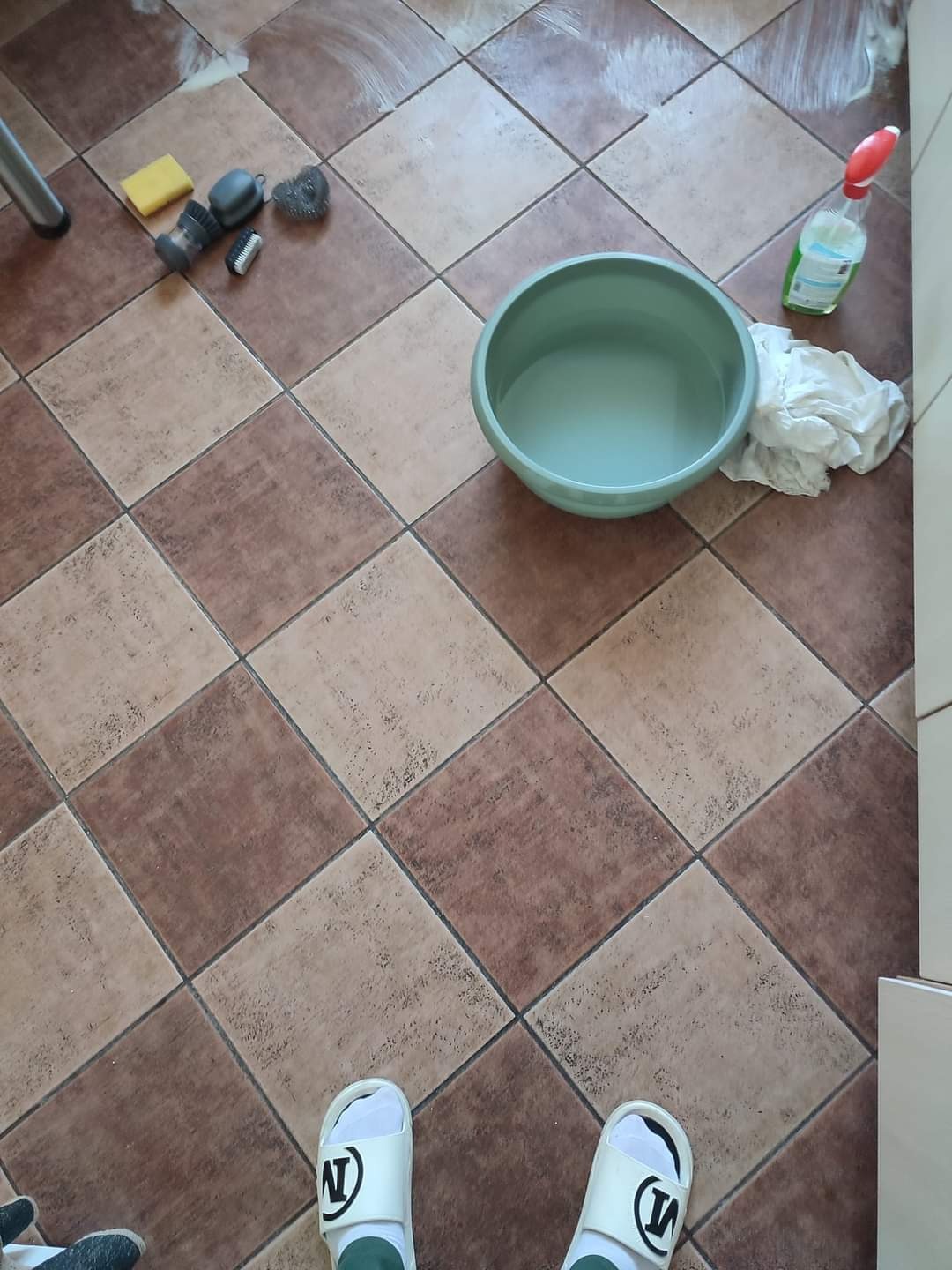 Ceramiczna podłoga w brązowo-beżową szachownicę z widocznymi śladami czyszczenia, miska z wodą, butelka ze spryskiwaczem, ściereczka, gąbka, szczotki i stopy osoby w białych klapkach.